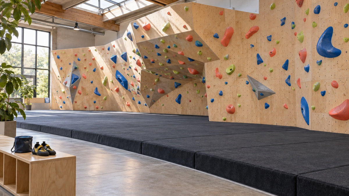 Empty bouldering gym with colorful holds, padded flooring, and climbing shoes and a chalk bag on a wooden bench.